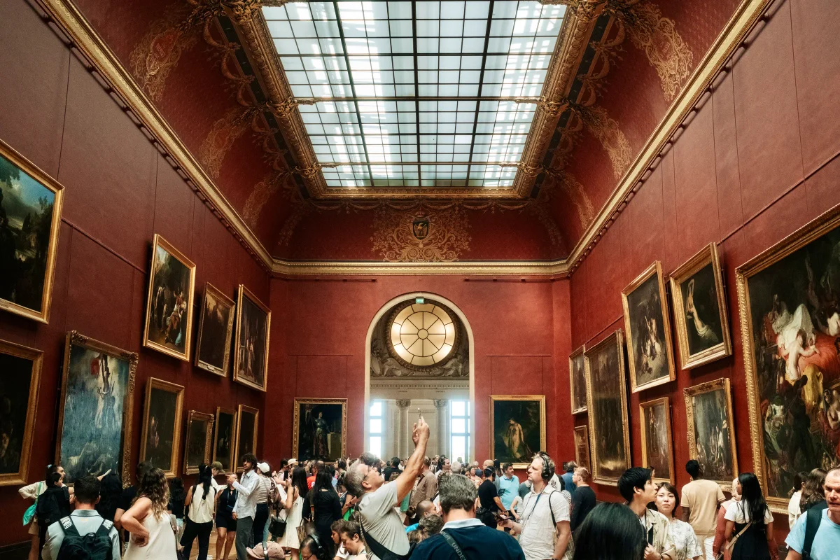 Crowds under the skylight of a red-walled gallery hung with paintings in the Louvre