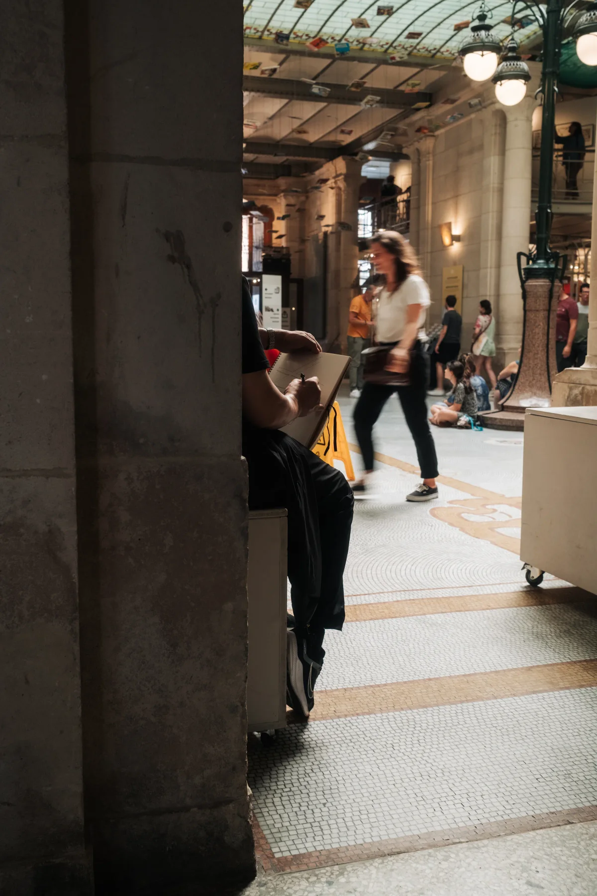 Artist sketching behind a stone pillar in a glass-roofed shopping arcade