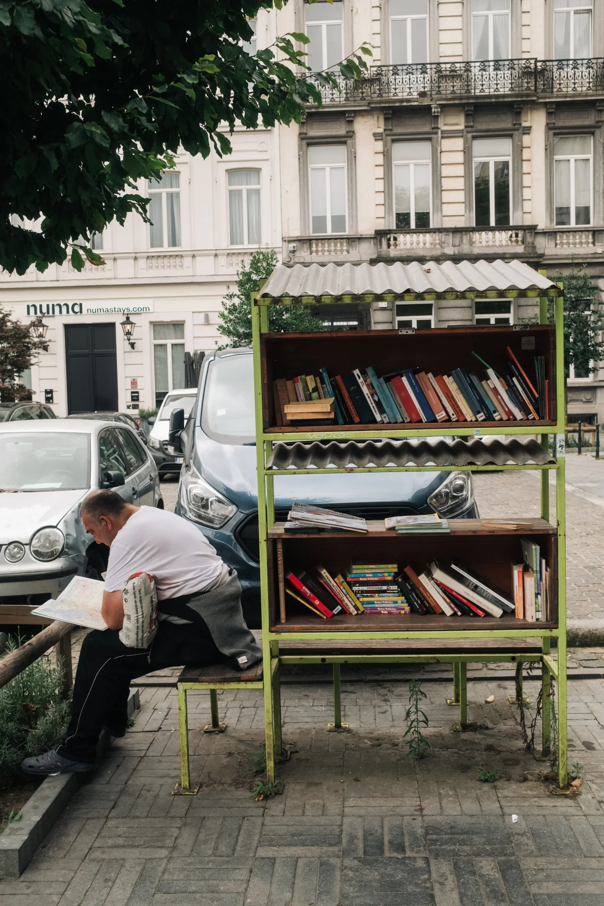Man reading beside a small outdoor book-exchange shelf on a city square