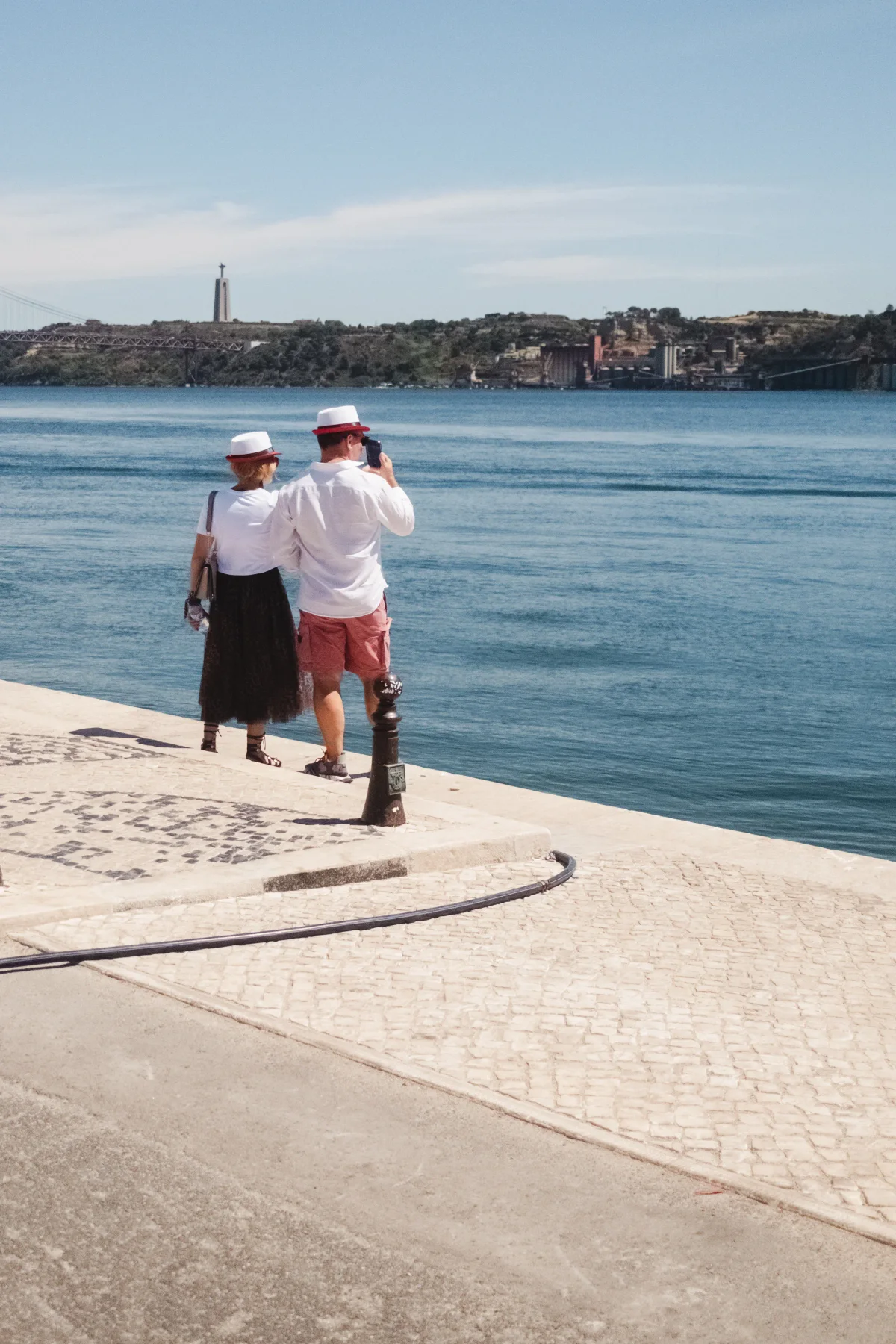 Older couple in matching white hats by the Tagus River with Cristo Rei across the water