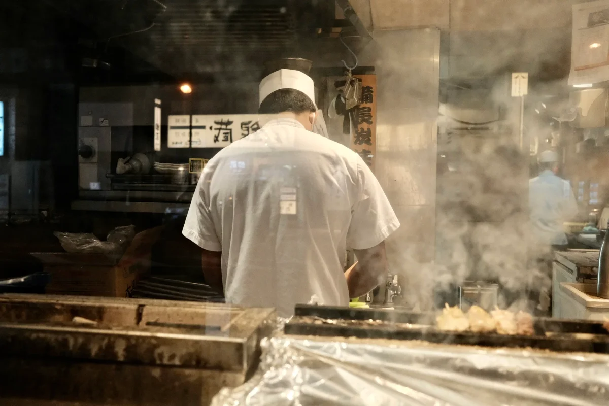 Cook in a white hat working at a smoking grill behind glass in a Japanese restaurant