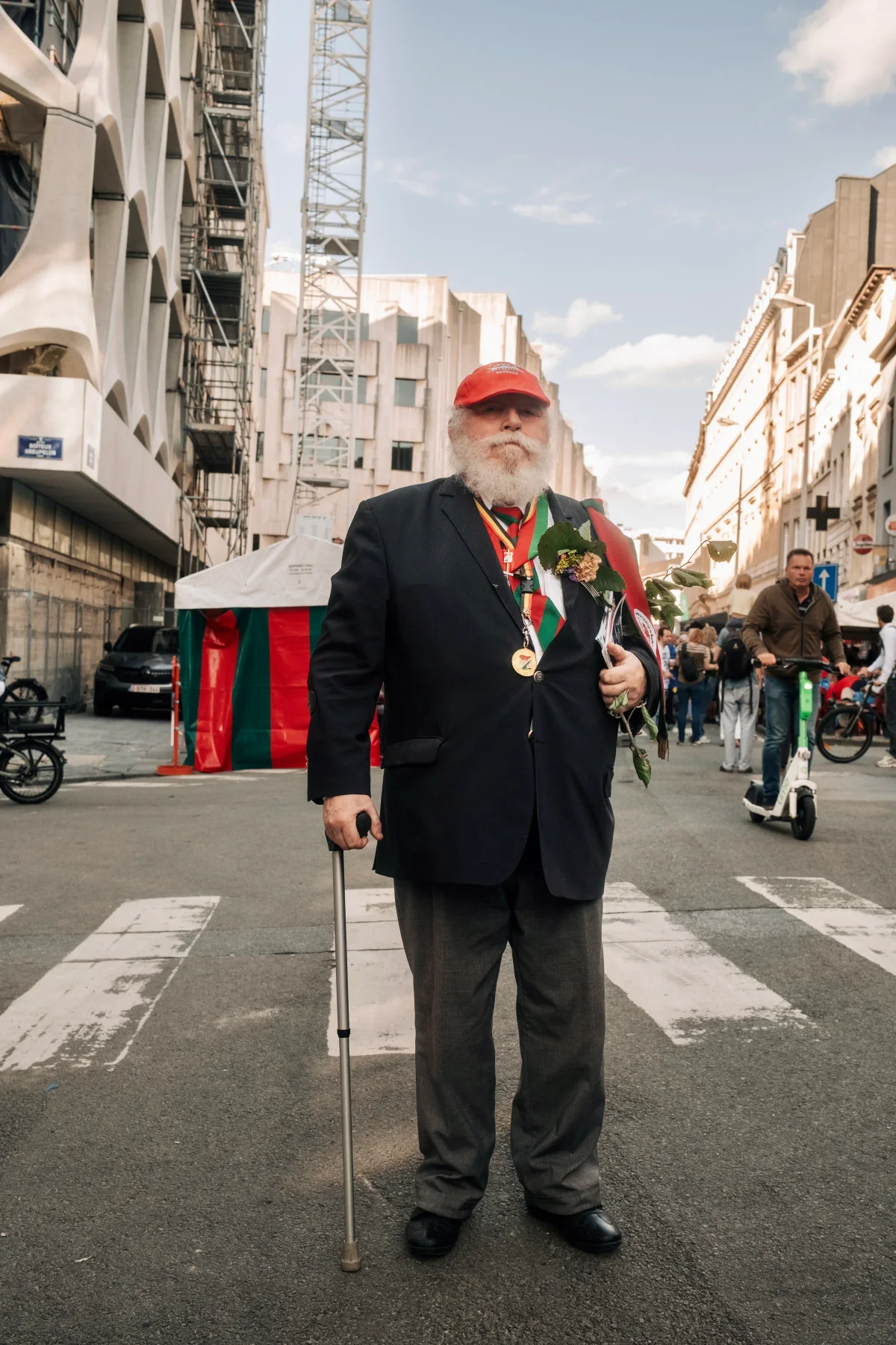 Elderly bearded man in a dark blazer, red cap and medals standing with a cane on a city street