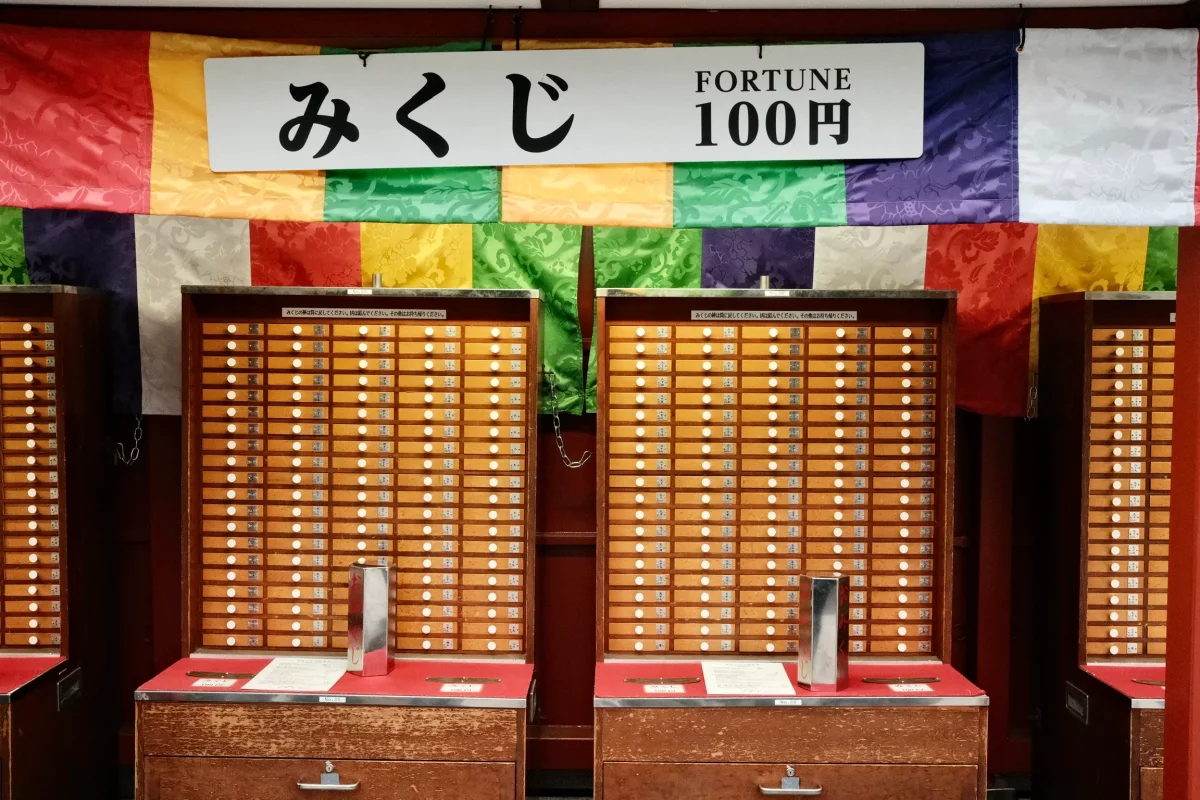Wooden omikuji fortune drawers beneath a sign reading Fortune 100 yen and colorful silk banners