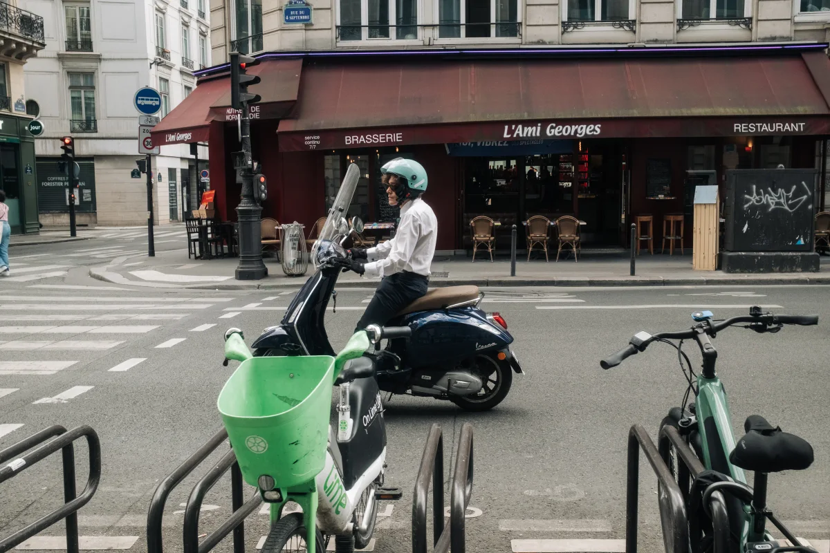 Rider on a navy Vespa passing a brasserie as bikes stand in a rack