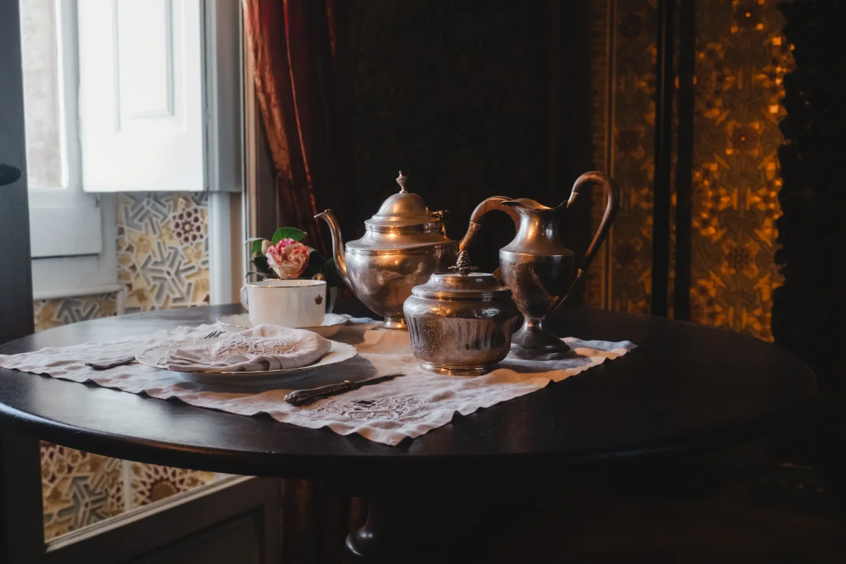 Silver teapot and tea service on a round table beside a tiled window