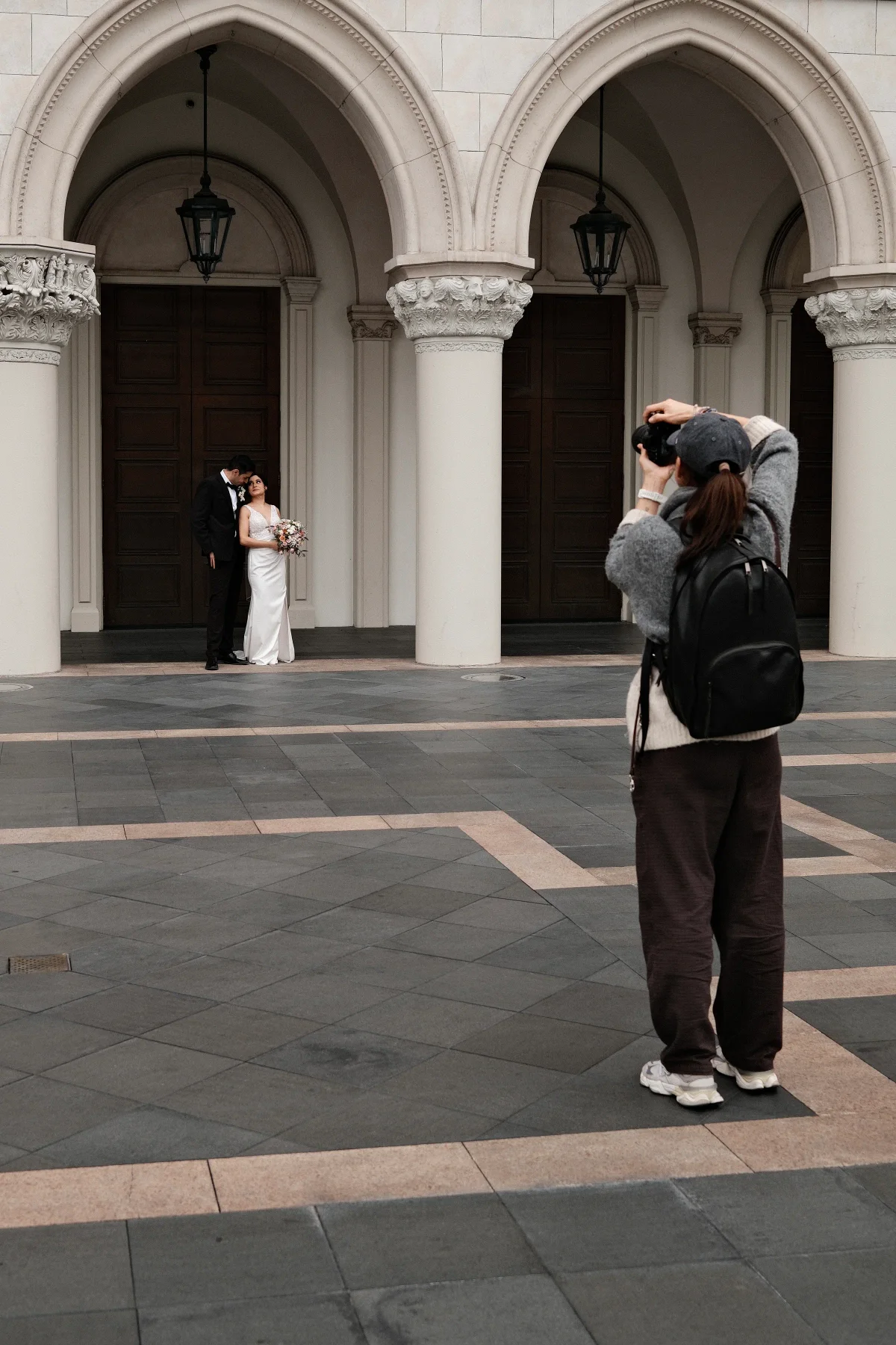 Photographer in a backpack shooting a bride and groom beneath a white stone arcade