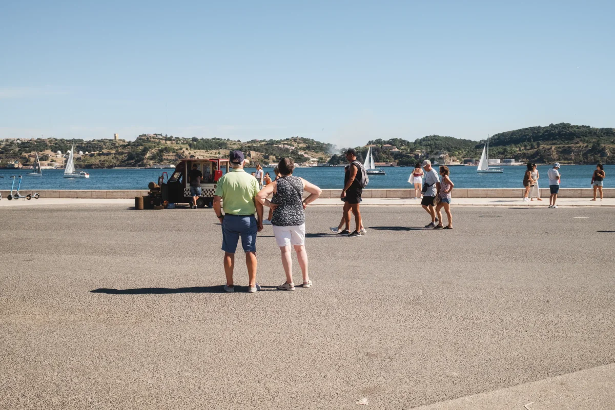 Couple and tourists on a sunny riverside promenade with sailboats on the water