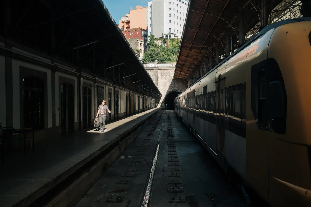 Man walking a sunlit platform beside a train in a covered station with a tunnel ahead