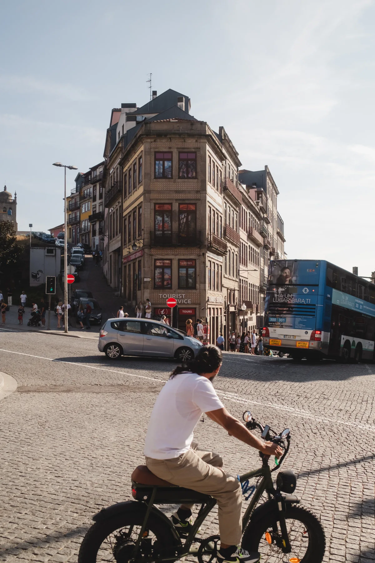 Man riding an electric bike across a cobblestone square past old stone buildings and a tour bus