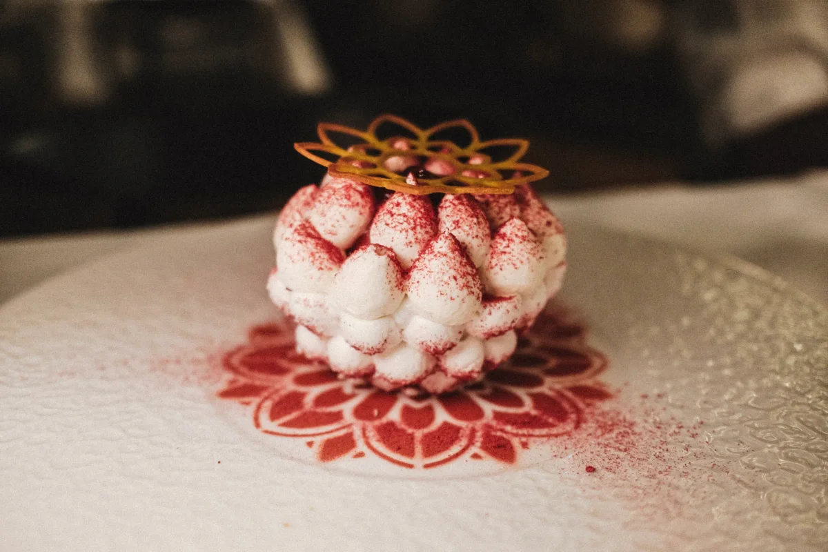 Plated white meringue dessert topped with a lace sugar crown and dusted red powder