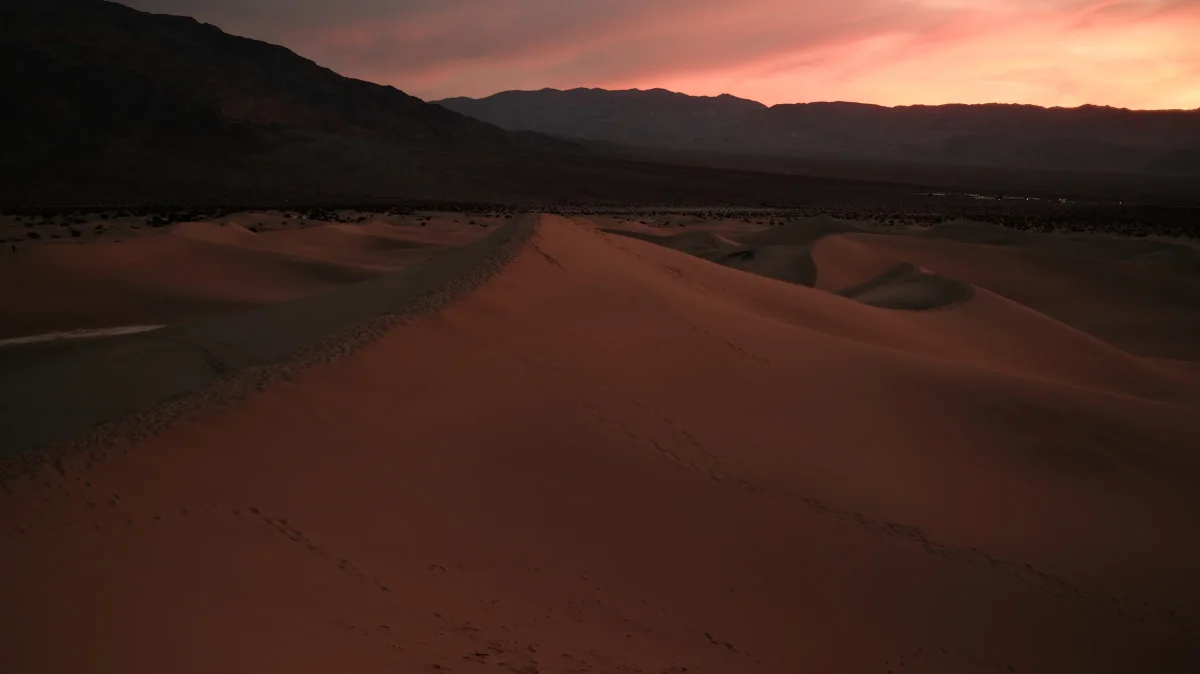 Curving sand dune ridge at twilight beneath a pink sky and dark mountains