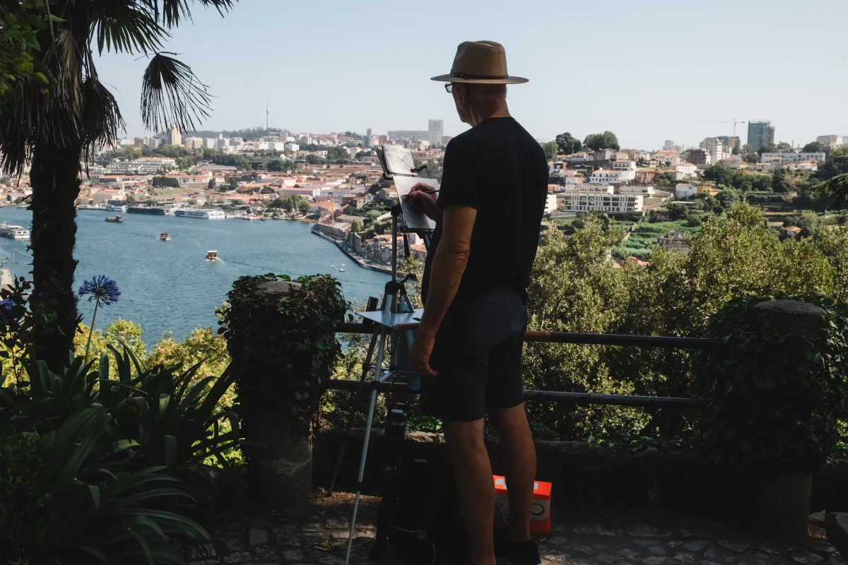 Man in a straw hat painting at an easel overlooking a river city