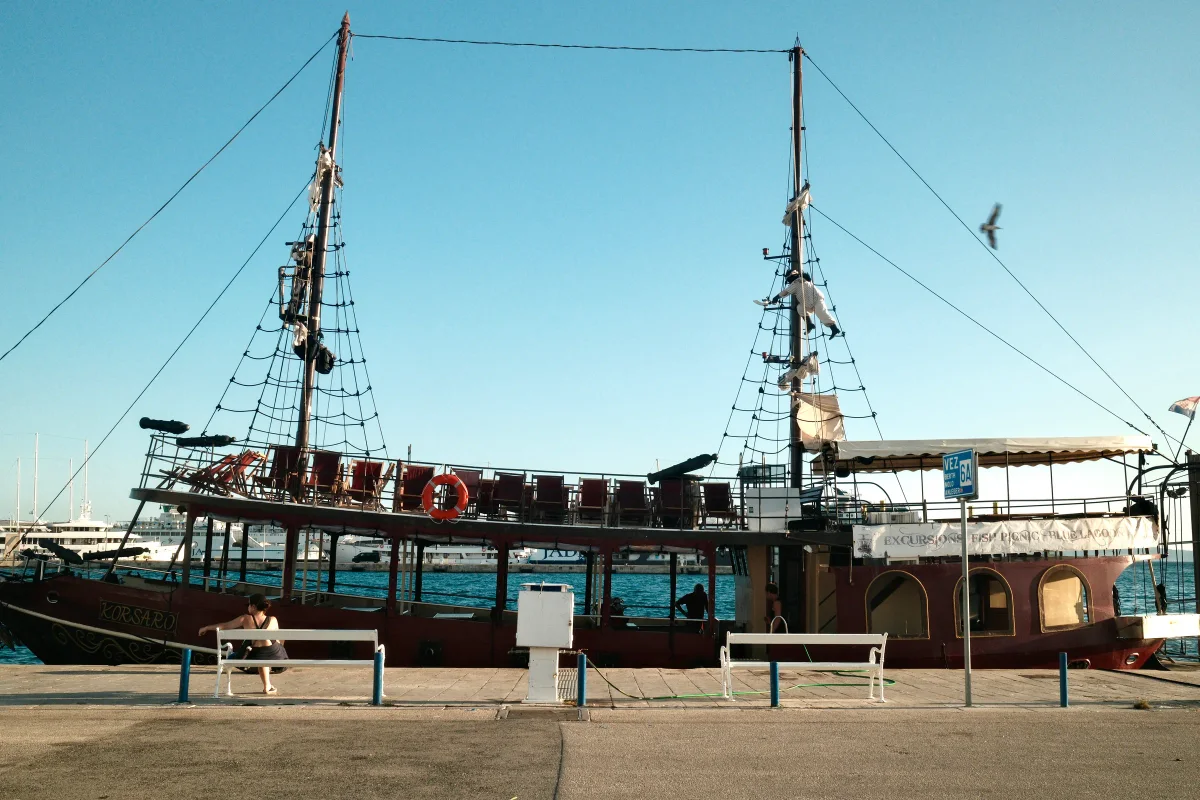 Two-masted wooden tour ship moored at a harbor promenade under a blue sky