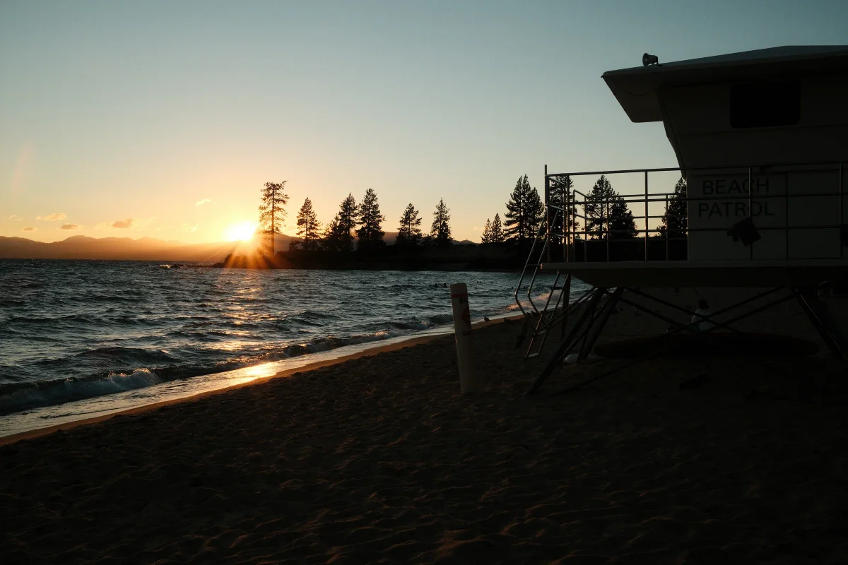 Sun setting behind pine trees over a lake beach, with a beach patrol tower in silhouette