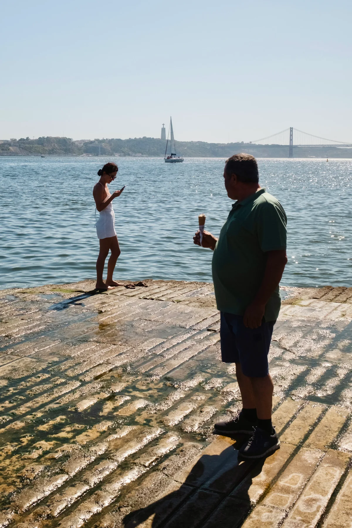 Man with an ice cream and a woman in white on a wet ramp before the 25 de Abril Bridge