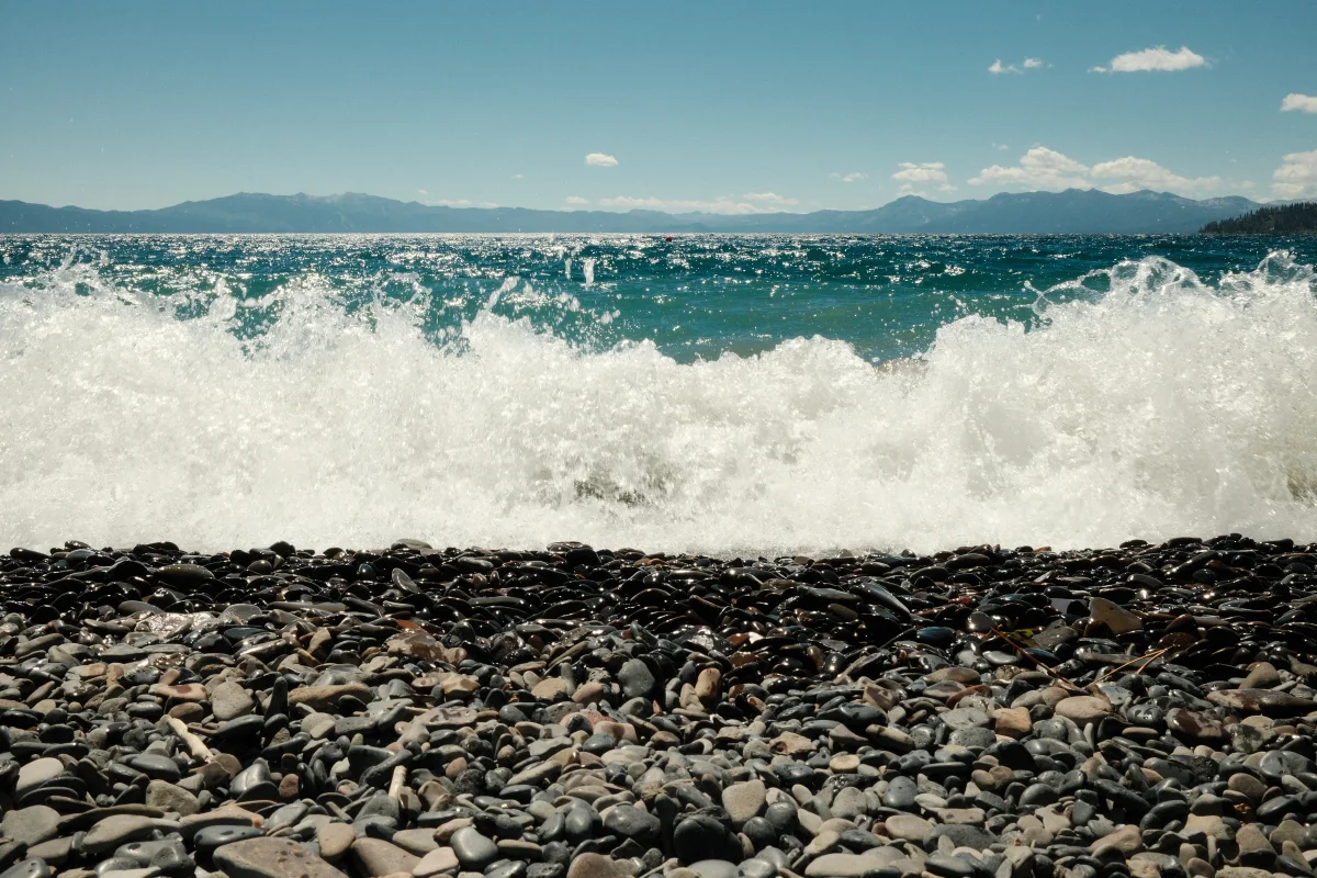 White wave crashing onto a pebble shore with blue water and distant mountains