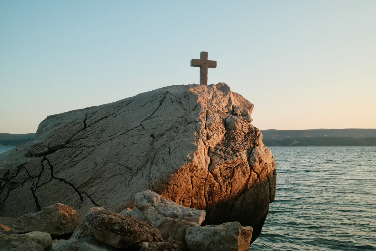 Stone cross atop a large sunlit boulder beside the sea