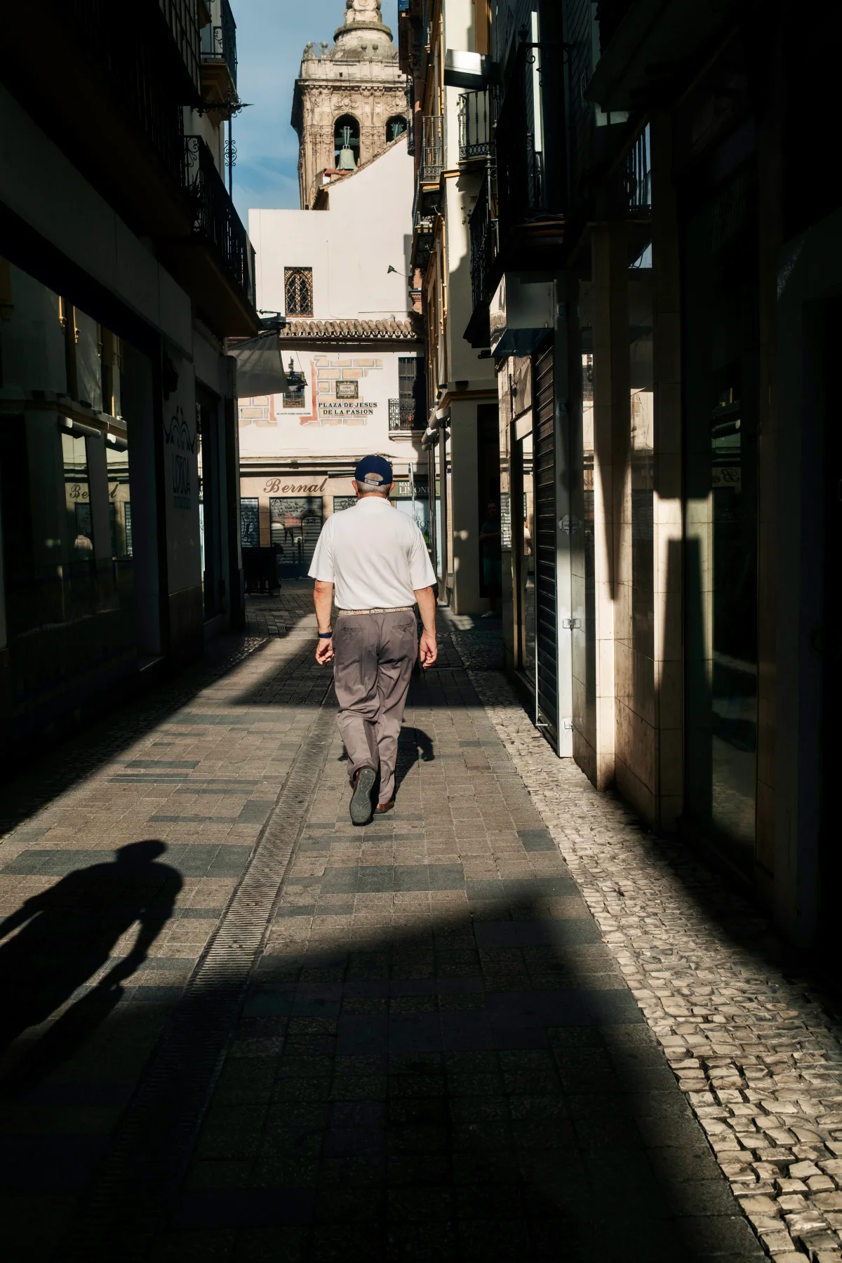 Man in a white shirt walking down a shaded street toward a church bell tower