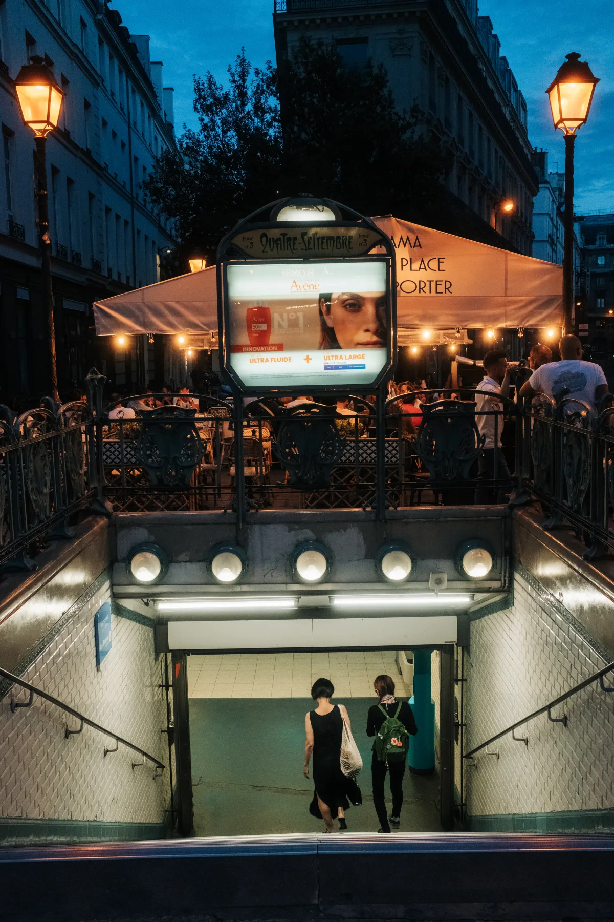 Quatre-Septembre Metro entrance at dusk with street lamps and a café terrace