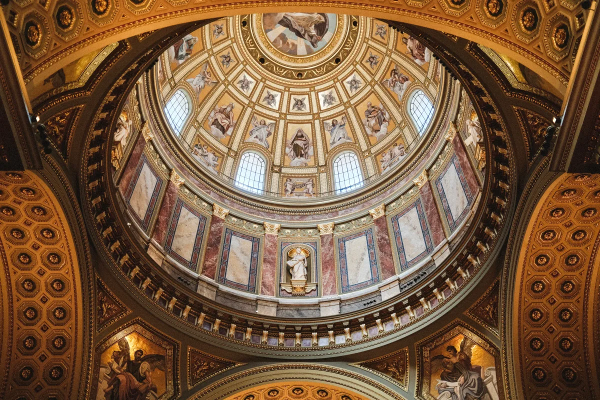 Looking straight up into an ornate gilded church dome with frescoed saints and arched windows
