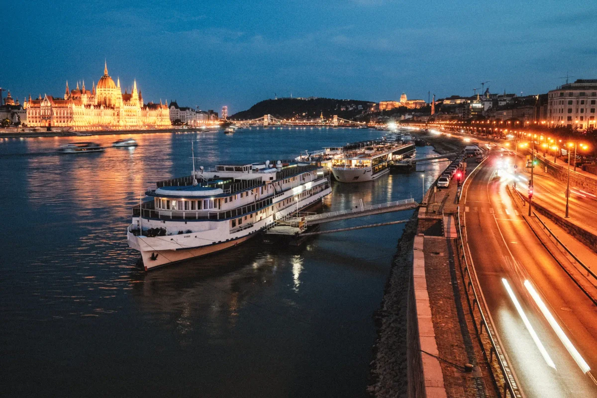Hungarian Parliament lit gold across the Danube at blue hour, with riverboats and car light trails